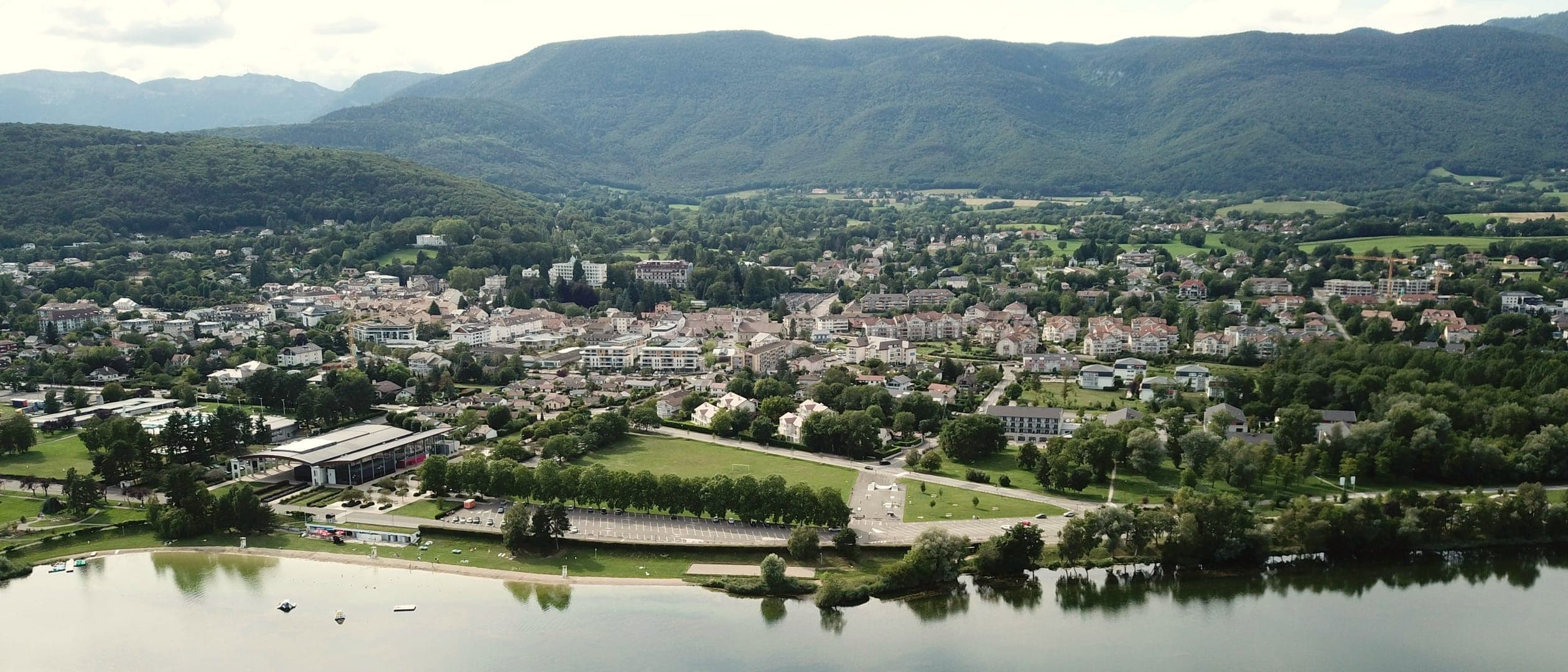 Vue aérienne d'une petite ville entourée de verdure au bord d'un lac, adossée à des montagnes boisées sous un ciel couvert.