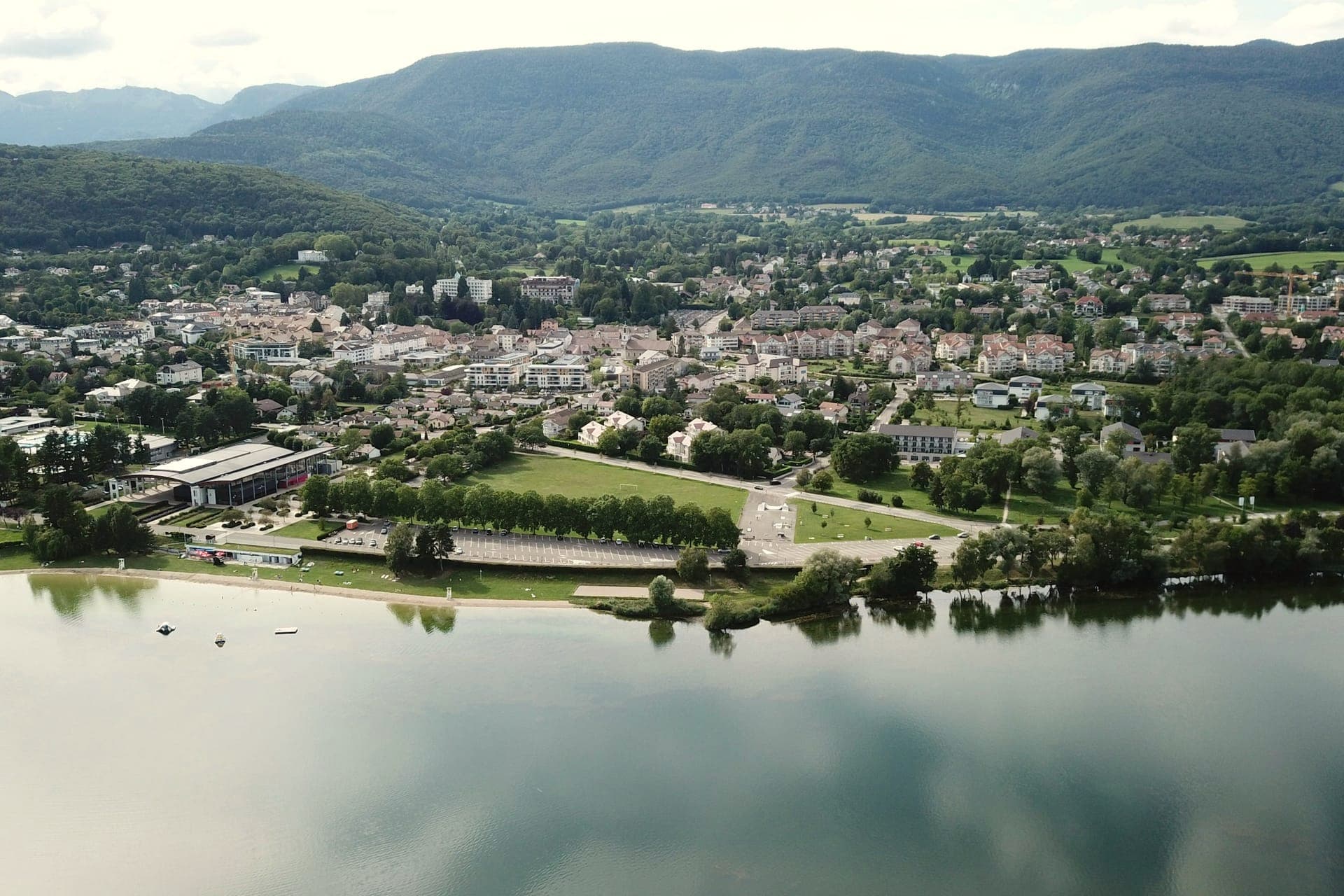 Vue aérienne d'une petite ville entourée de verdure au bord d'un lac, adossée à des montagnes boisées sous un ciel couvert.