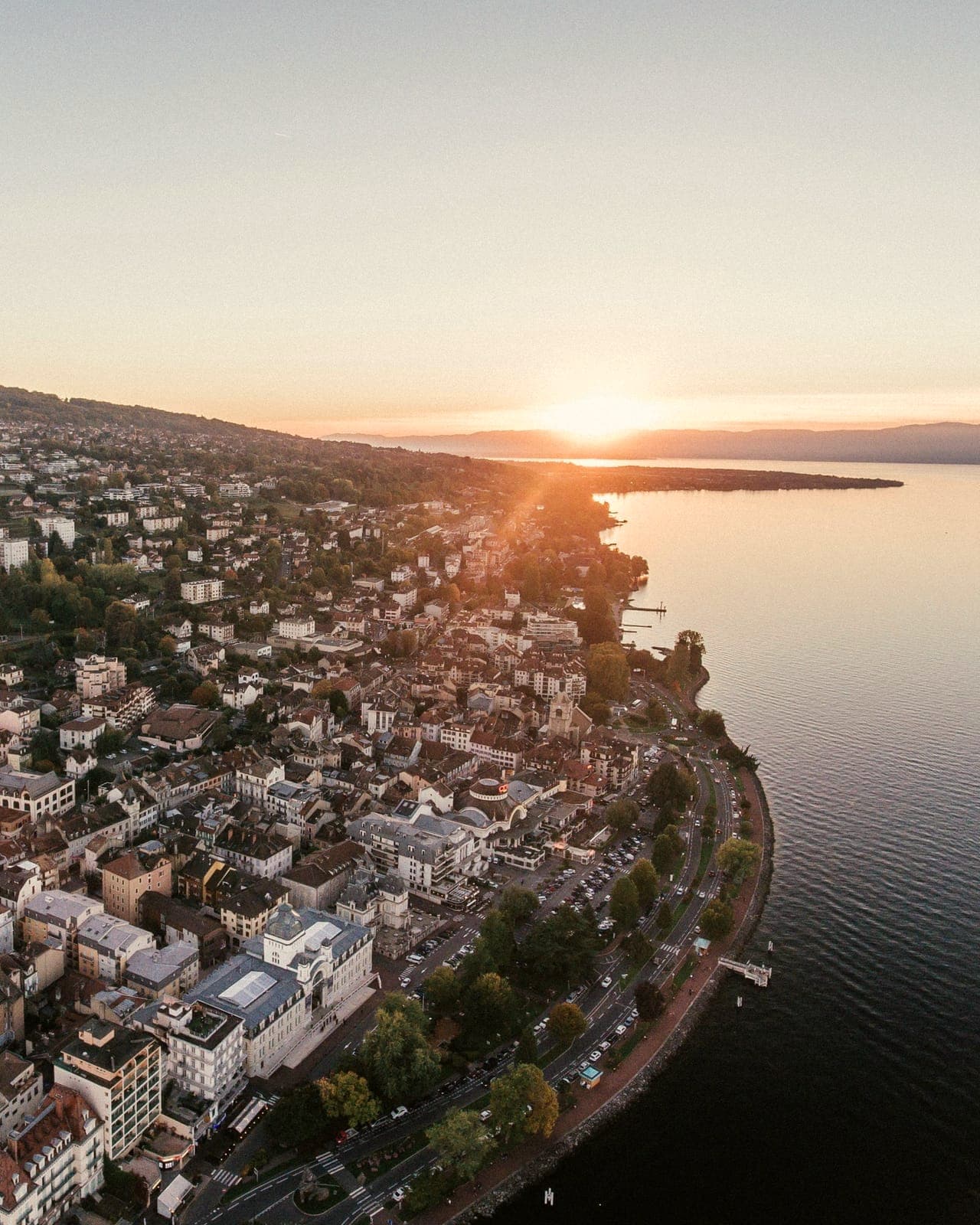Vue aérienne d'une ville au bord d'un lac au coucher du soleil : immeubles et toits au premier plan, quai arboré le long de l'eau, rive et collines à l'horizon.