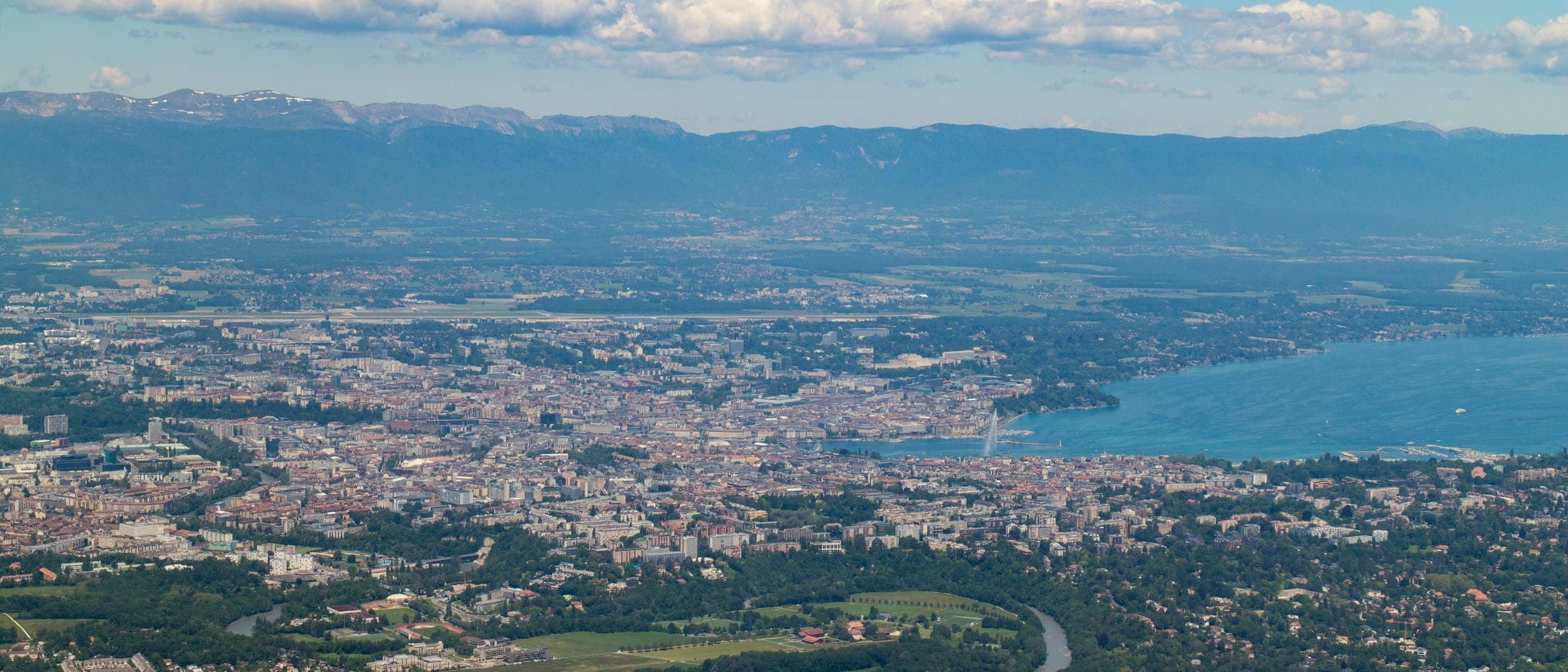 Vue plongeante sur une agglomération au bord d'un lac : champs et quartiers résidentiels au premier plan, une rivière en méandre, la ville et le jet d'eau, puis une longue chaîne de montagnes à l'horizon.