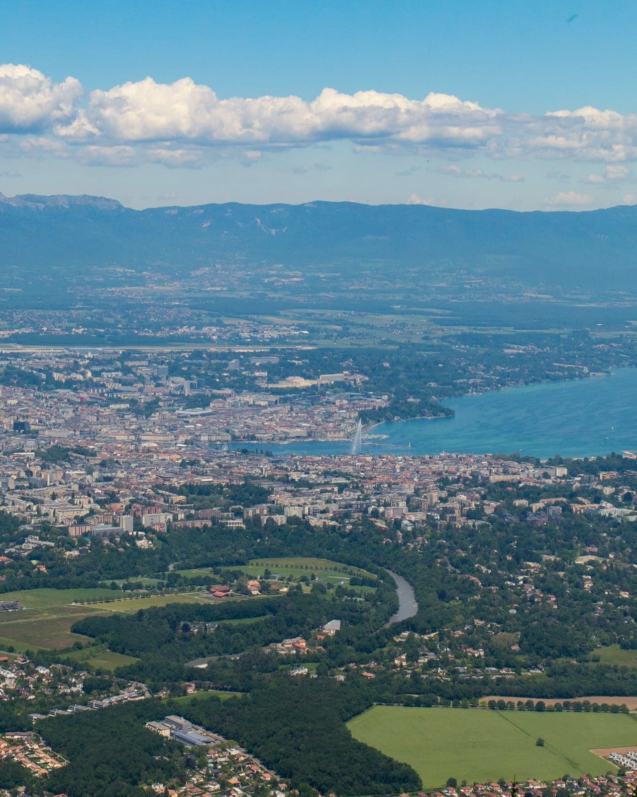 Vue plongeante sur une agglomération au bord d'un lac : champs et quartiers résidentiels au premier plan, une rivière en méandre, la ville et le jet d'eau, puis une longue chaîne de montagnes à l'horizon.