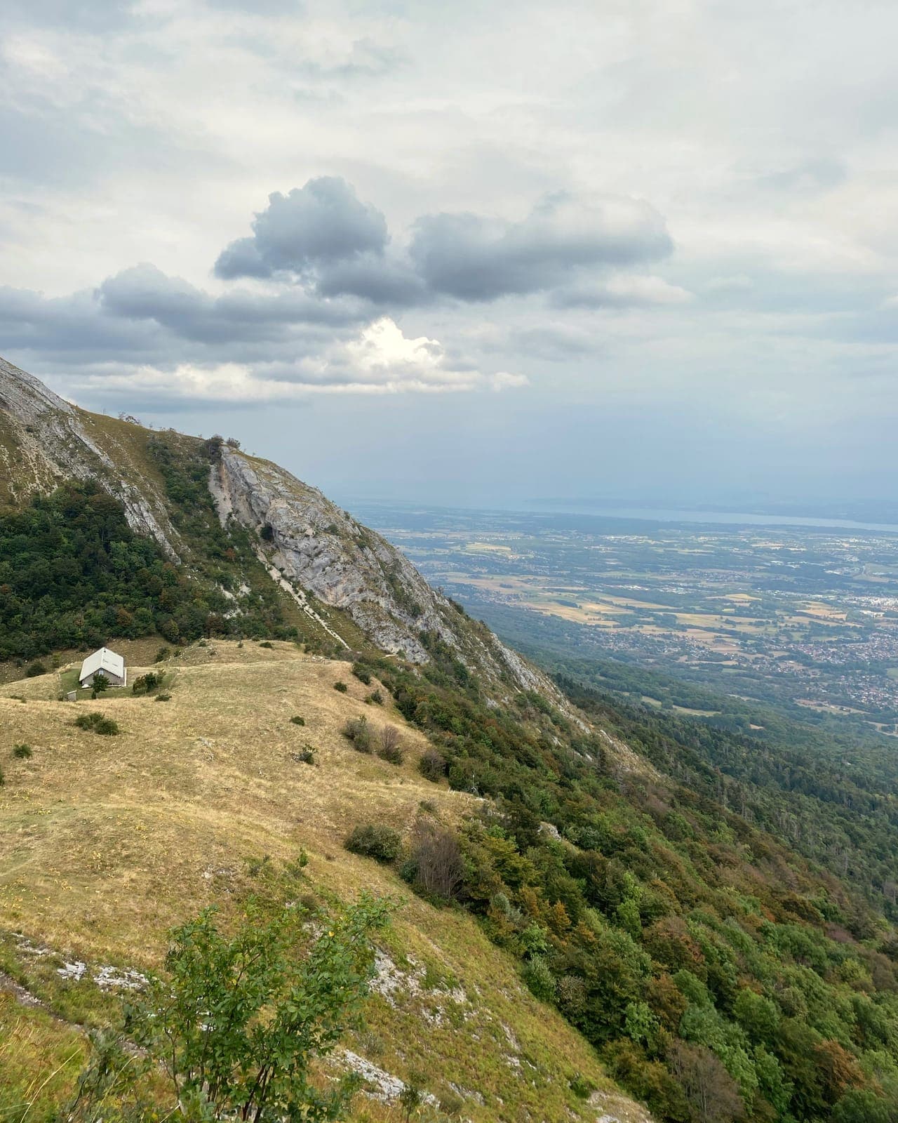 Pente herbeuse avec un petit chalet sous une crête rocheuse ; à droite, le versant boisé plonge vers une plaine parsemée de villages et un lac au loin, sous un ciel nuageux.