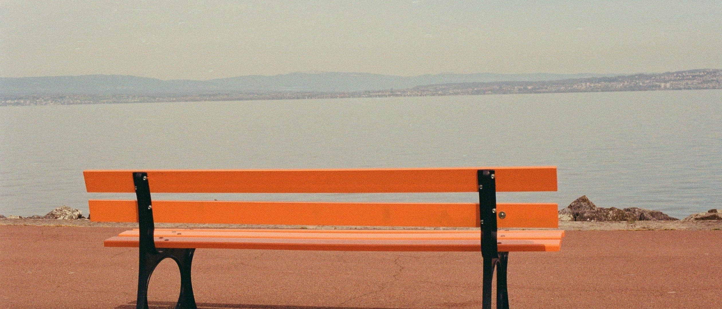 Un banc orange vide sur une promenade au bord du Léman ; au-delà, le lac calme et la rive opposée sous un ciel voilé.