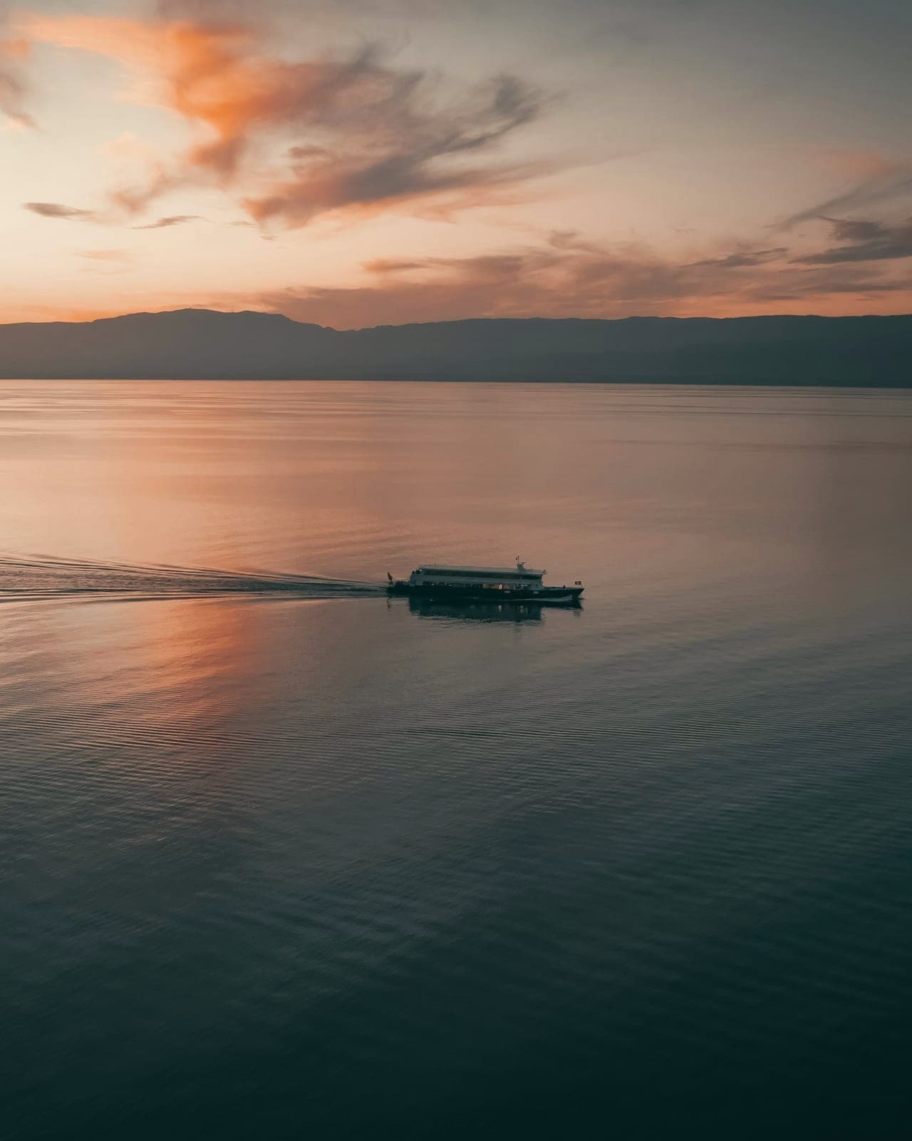 Vue en hauteur du Léman au crépuscule : un bateau traverse le lac calme en laissant un sillage, sous un ciel rose et gris, avec une ligne de collines sombres à l'horizon.
