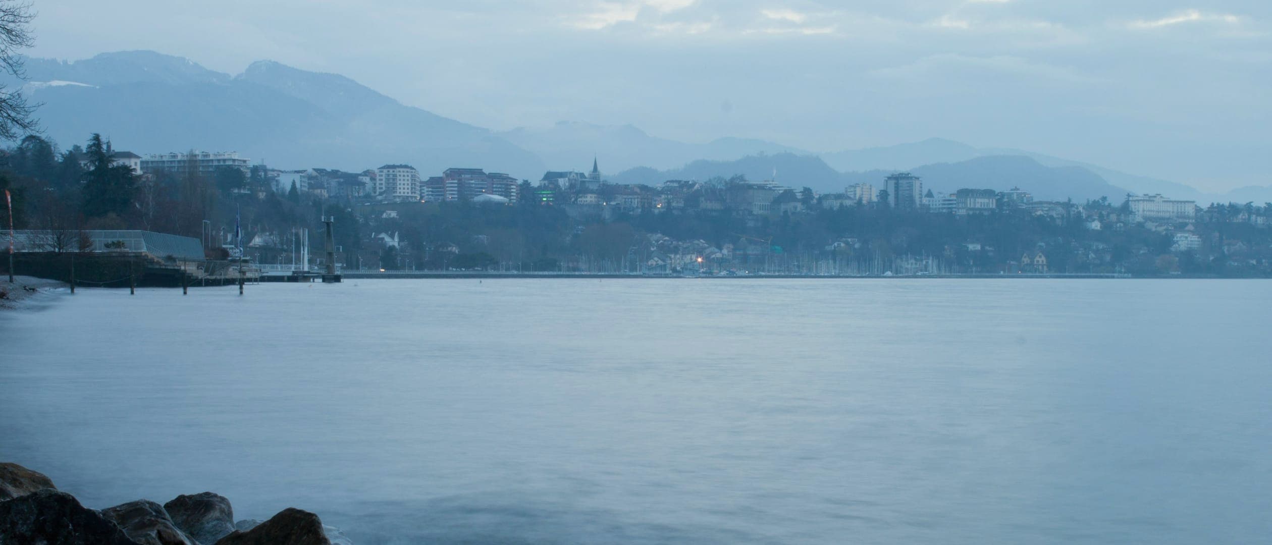 Rochers au premier plan au bord d'un lac lisse aux tons bleus ; en face, une ville étagée sur la rive et des montagnes dans la brume.