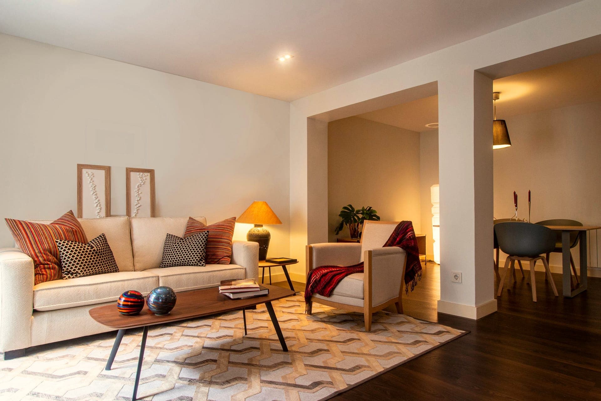 Un salon d'appartement en lumière chaude : canapé clair, coussins, lampe allumée et table basse.