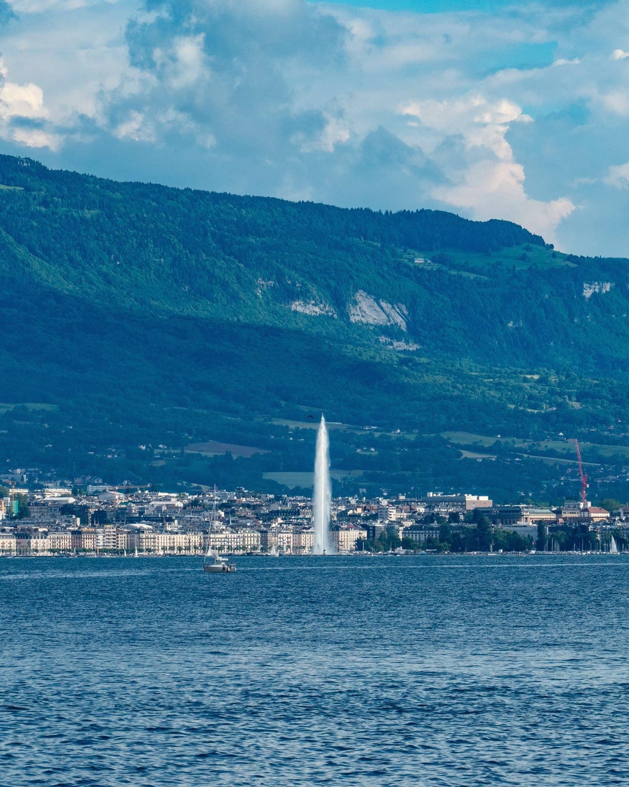 Le jet d'eau de Genève s'élève devant une rangée d'immeubles au bord du lac ; derrière, une longue montagne boisée aux falaises claires, sous un ciel nuageux.
